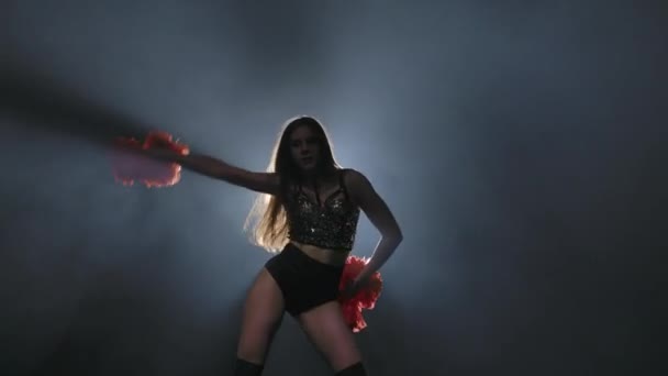 Young female cheerleader with orange pompoms in uniform is dancing on black smoky studio background with backlight. Silhouette of dancer performs cheering dance. Close up. Slow motion ready 59.94fps. — Stockvideo