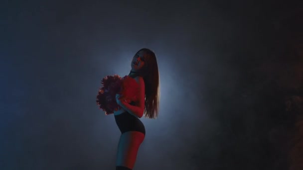 Giovane cheerleader femminile con pompon arancioni in uniforme sta ballando su sfondo nero fumoso studio con retroilluminazione. La silhouette di ballerino compie la danza allegra. Chiudete. Movimento lento pronto 59.94fps. — Video Stock