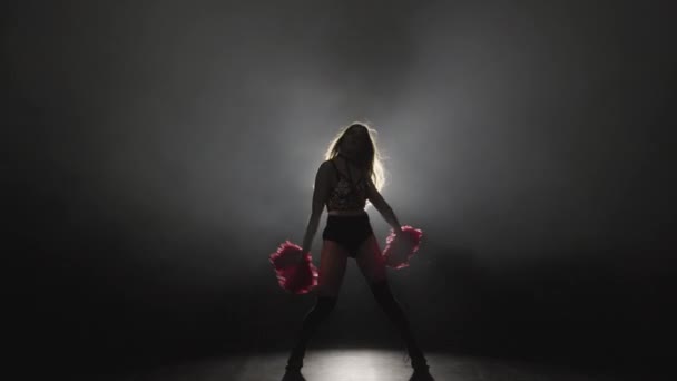 Jonge vrouwelijke cheerleader met oranje pompons in uniform danst op zwarte rokerige studio achtergrond met achtergrondverlichting. Silhouet van mooie danseres vertolkt juichende dans. Slow motion klaar 59,94fps. — Stockvideo