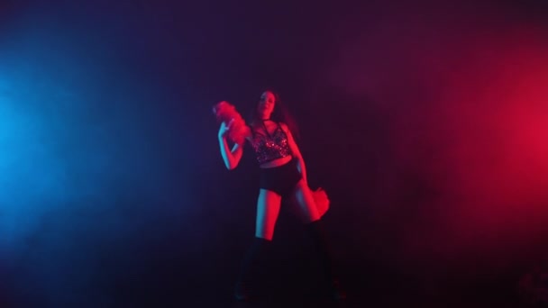 Young cheerleader with pompoms in uniform is dancing on black smoky studio background with red and blue lights. Silhouette of beautiful dancer performs cheering dance. Slow motion ready 59.94fps. — Stockvideo