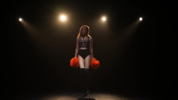 En söt cheerleader med pompoms dansar på en svart studio bakgrund med ljus och rök. Ung kvinna viftar med händerna och flyttar höfterna. Slow motion klar 59,94fps. — Stockvideo