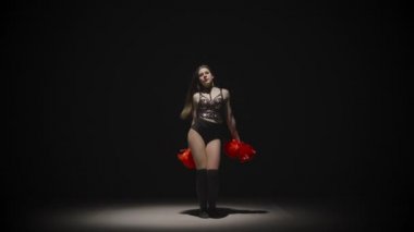 Uzun saçlı, parlak saçlı ponpon kız güçlü bir şekilde dans eder ve ponponlarını sallar. İnce kadın dansçı ellerini ve kalçalarını arka plandaki siyah stüdyoda hareket ettiriyor. Yavaş çekim hazır 59.94fps.