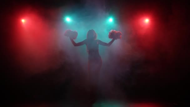 Silhueta de líder de torcida delgado dançando com pompons contra um fundo de fumaça e luzes azuis e vermelhas brilhantes em um estúdio escuro. Líder de torcida, dança de torcida, fitness. Movimento lento pronto 59.94fps. — Vídeo de Stock