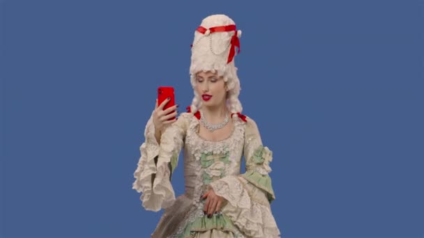 Porträt einer Höflichkeitsdame in weißem Vintage-Spitzenkleid und Perücke, die sich per Handy für einen Videochat unterhält. Junge Frau posiert im Studio mit blauem Hintergrund. Aus nächster Nähe. Zeitlupe bereit 59.94fps.