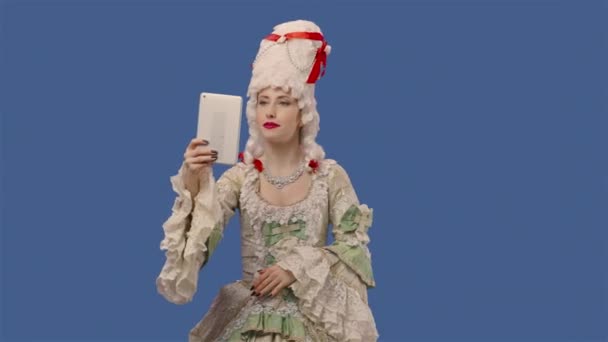 Portrait of courtier lady in white vintage dress and wig talking for video chat using digital tablet. Young woman posing in studio with blue screen background. Close up. Slow motion ready 59.94fps. — стоковое видео