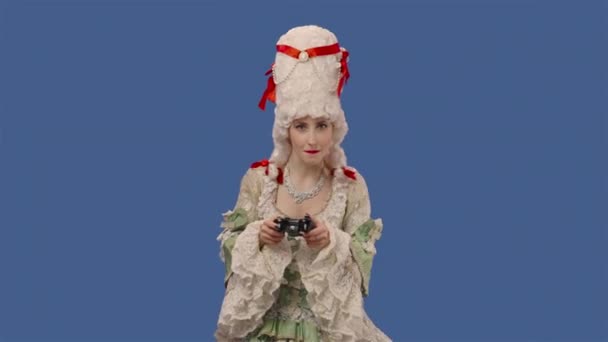 Portrait de courtisane en robe vintage et perruque jouer à un jeu vidéo à l'aide d'une manette sans fil, perd. Jeune femme posant en studio avec fond bleu. Ferme là. Ralenti prêt 59.94fps. — Video