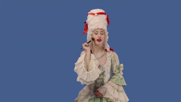 Retrato de dama cortesana en vestido vintage y peluca está aplicando polvo a su cara usando un cepillo de maquillaje. Mujer joven posando en el estudio con fondo de pantalla azul. De cerca. Cámara lenta lista 59.94fps. — Vídeo de stock