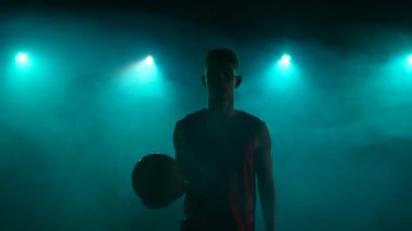 Kameraya bakıp topu fırlatan genç basketbolcunun portresi. Spor kıyafetli bir sporcu. Stüdyonun arka planında duman ve mavi ışıklarla poz veriyor. Kapatın. Yavaş çekim hazır 59.94fps.