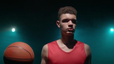 Elinde top olan genç bir basketbolcunun portresi. Kırmızı spor üniformalı bir atlet. Stüdyonun arka planında dumanlı ve mavi ışıklarla poz veriyor. Kapatın. Yavaş çekim hazır 59.94fps.