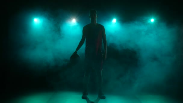 Silhueta de um jovem rapaz driblando basquete em um fundo escuro estúdio com fumaça e luzes azuis. Jovem desportista alto de uniforme desportivo bate bola no chão. Movimento lento pronto 59.94fps — Vídeo de Stock