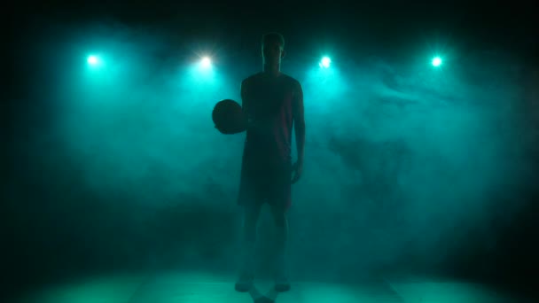 Silhueta de um jovem jogador de basquete jogando a bola para cima. Jovem desportista alto em uniforme desportivo posando em um fundo escuro estúdio com fumaça e luzes azuis. Movimento lento pronto 59.94fps. — Vídeo de Stock