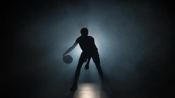 Silhouette of basketball player dribbles ball practicing skills in a dark studio with smoke and backlight. Professional player trains before basketball competition. Slow motion ready 59.94fps. — Stock Video