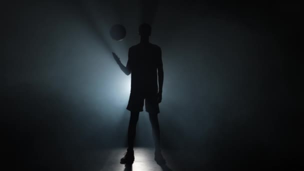 Silhouet van een jonge basketbalspeler die de bal omhoog gooit. Lange jonge sportman in sportuniform poserend op een donkere studio achtergrond met rook en achtergrondverlichting. Slow motion klaar 59,94fps. — Stockvideo