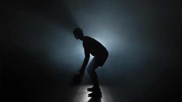 Silhouet van basketbalspeler dribbelt bal oefenen vaardigheden in donkere studio met rook en achtergrondverlichting. Professionele speler traint voor basketbal competitie. Zijaanzicht. Slow motion klaar 59,94fps — Stockvideo