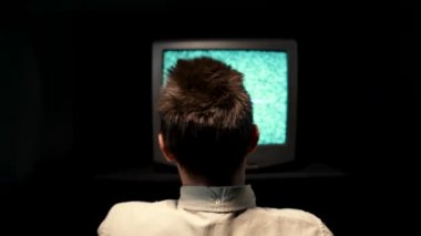 Arka planda, karanlık bir stüdyoda, eski bir televizyonun önünde oturan bir adam var. Siyah bir arka planda, televizyon ekranında parazit var. Adam kırık TV 'ye elini vurdu. Kapat..
