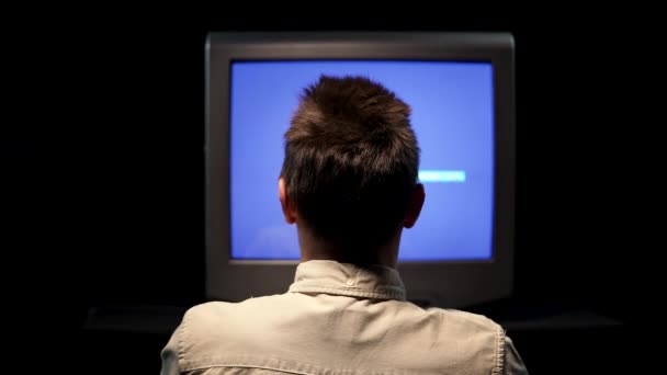 Rückansicht eines Mannes, der vor einem alten Fernseher mit Störgeräuschen auf einem blauen TV-Monitor in einem dunklen Studio vor schwarzem Hintergrund sitzt. Der Mann klopft mit der Hand auf den kaputten Fernseher. Nahaufnahme. — Stockvideo