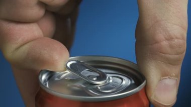 Erkek el, mavi arka planda sallanmış bir bira kutusunu açıyor. Bir kutu karbonatlı içecekten köpük çeşmesi dökülüyor. Ağır çekim. Kapat..
