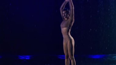 Hafif tefekkür, yağmurda esnek bir bayan dansçı tarafından koyu renkli arka planda mavi ışıklarla sunuluyor. Genç ıslak yarı çıplak kadın yağmur damlaları arasında modern koreografi dansı yapıyor. Yavaş çekim.