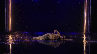 Hafif tefekkür, yağmurda esnek bir bayan dansçı tarafından koyu renkli arka planda mavi ışıklarla sunuluyor. Genç ıslak yarı çıplak kadın yağmur damlaları arasında modern koreografi dansı yapıyor. Yavaş çekim.