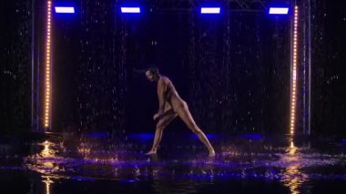 Hafif tefekkür, yağmurda esnek bir bayan dansçı tarafından koyu renkli arka planda mavi ışıklarla sunuluyor. Genç ıslak yarı çıplak kadın yağmur damlaları arasında modern koreografi dansı yapıyor. Yavaş çekim.