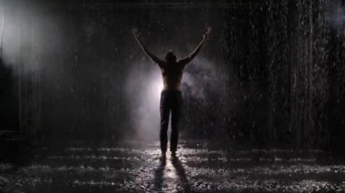 Modern çağdaş koreografinin duygusal performansı parlak spot ışıklarına karşı yağmurda. Duman şişeleri, erkek dansçının çıplak gövdesini su damlalarının arasına gizliyor. Yavaş çekim.