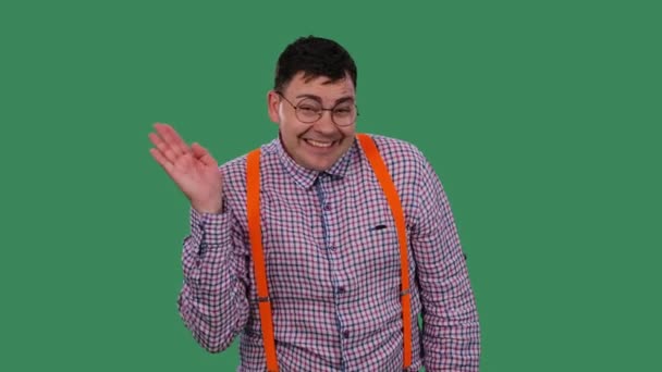 Porträtt av en leende man som tittar på kameran och viftar med handen i hälsning. En man i glasögon, i rutig skjorta med orange hängslen i studion på en grön skärm. Sakta i backarna. Närbild. — Stockvideo