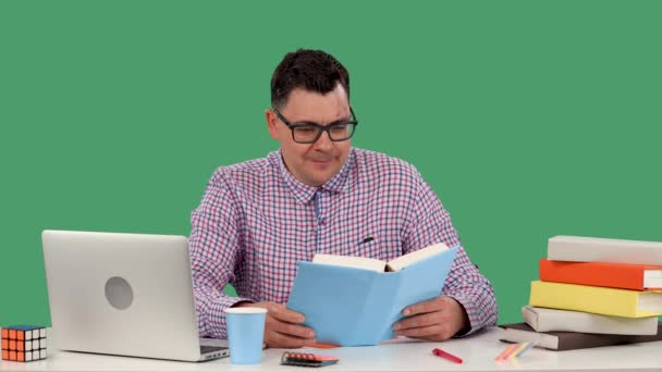A man in glasses sits at a desk in front of a laptop and reads a book, laughs, covering his mouth with his palm. A man in the studio on a green screen. Quality leisure. Slow motion. Close up. — Stock Video