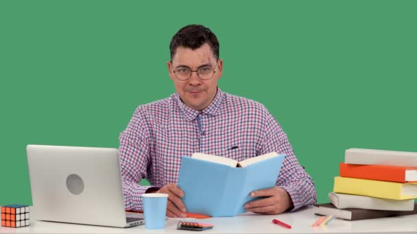 Un homme avec des lunettes est assis à un bureau devant un ordinateur portable et lit un livre, sourit, pose son poing sur son menton, complote quelque chose. Portrait d'un homme en studio sur un écran vert. Au ralenti. Gros plan — Video