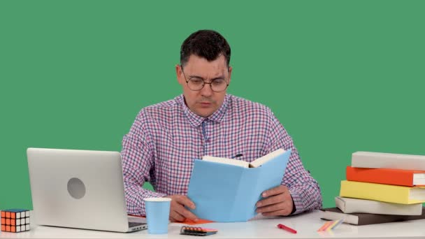 Un hombre con gafas se sienta en un escritorio frente a un portátil y lee un libro, se estremece, se pone el puño en la barbilla, preguntándose qué ha leído. Un hombre en el estudio en una pantalla verde. En cámara lenta. De cerca.. — Vídeos de Stock