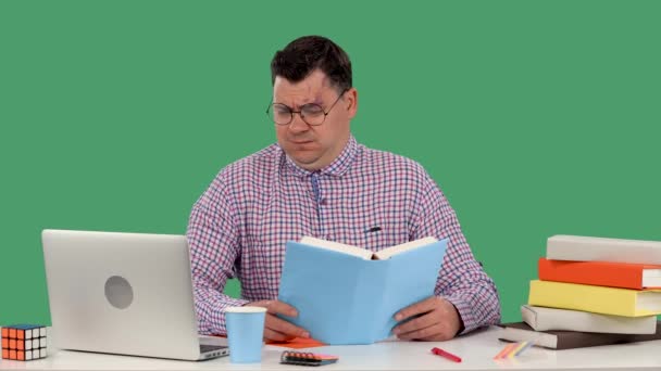 Um homem com óculos senta-se em uma mesa na frente de um laptop e lê um livro, acena com a cabeça negativamente, fica desagradavelmente surpreso, chocado. Um homem no estúdio num ecrã verde. Movimento lento. Fechar. — Vídeo de Stock