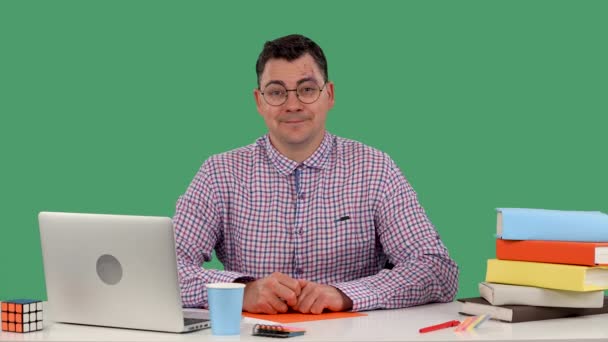 一个戴眼镜的男人坐在笔记本电脑前的书桌前，直视四周，带着神秘的微笑把眼镜挺直。一个男人在工作室的肖像在一个绿色的屏幕上。慢动作靠近点. — 图库视频影像
