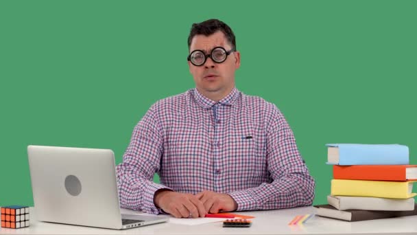 Un hombre se sienta en un escritorio frente a una computadora portátil y libros, piensa en algo, asiente con la cabeza negativamente, no hay ideas. Un hombre con gafas en el estudio en una pantalla verde. En cámara lenta. De cerca.. — Vídeo de stock