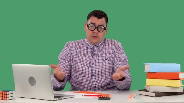 A disgruntled man sits at a desk in front of a laptop and books, points his hands in front of him, indignant. Portrait of a man with glasses in the studio on a green screen. Slow motion. Close up. — Stock Video