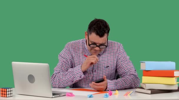 En man gör anteckningar i en anteckningsbok, höjer handen med en penna upp, har en idé. En man poserar medan han sitter vid ett skrivbord framför en bärbar dator och böcker i studion på en grön skärm. Sakta i backarna. Närbild. — Stockvideo