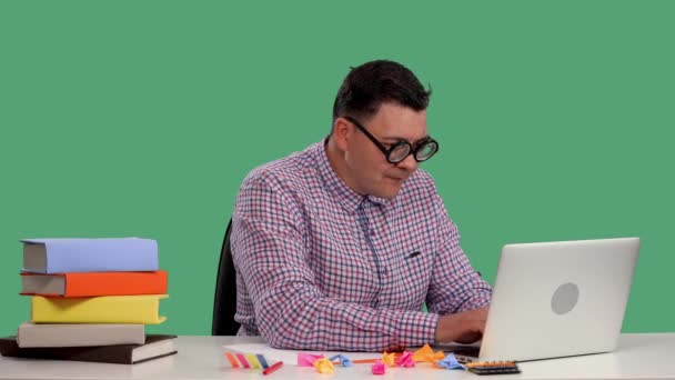 Un homme s'assoit à une table, utilise un ordinateur portable, tape sur un clavier, rit, se couvre la bouche avec sa paume et regarde autour de lui. Portrait d'un homme en studio sur un écran vert. Au ralenti. Gros plan. — Video