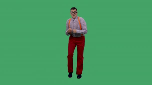 Un retrato de un hombre bailando y cantando junto a la música. Un hombre en gafas con lentes gruesas, en una camisa a cuadros con tirantes anaranjados en el estudio en una pantalla verde. Movimiento lento. — Vídeos de Stock