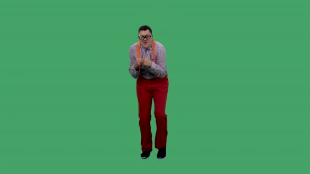 El hombre, encorvado, baila y canta junto a las melodías. Retrato de un hombre en gafas con lentes gruesas, en una camisa a cuadros con tirantes anaranjados en el estudio en una pantalla verde. Movimiento lento. — Vídeos de Stock