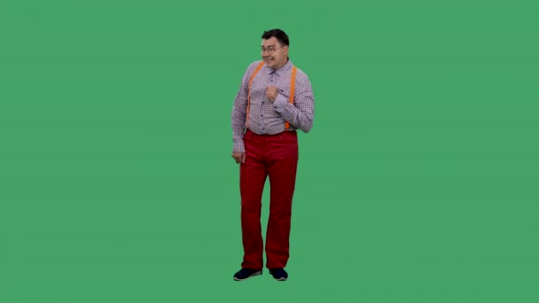Een man met een sluwe blik, luistert naar iets en wiebelt met zijn wenkbrauwen. Portret van een man in een bril, in een shirt met oranje bretels in de studio op een groen scherm. Langzame beweging. — Stockvideo