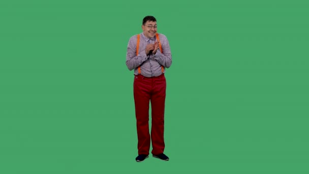 Uśmiechnięty mężczyzna drapie się po dłoni. Portret mężczyzny w okularach, w koszuli w kratę z pomarańczowymi szelkami w studiu na zielonym ekranie. Zwolniony ruch. — Wideo stockowe