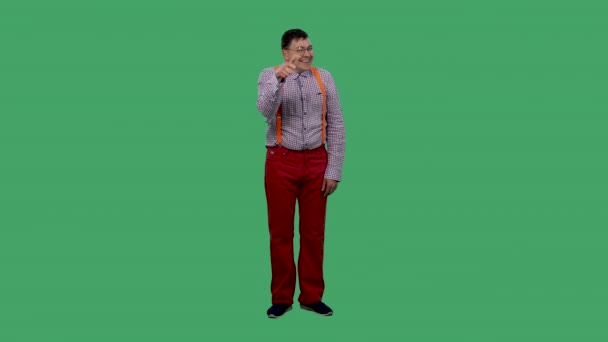 微笑的男人用他的食指指着摄像机说你你你一个戴眼镜的男人，穿着格子衬衫，在工作室的绿色屏幕上挂着橙色的吊带。慢动作. — 图库视频影像