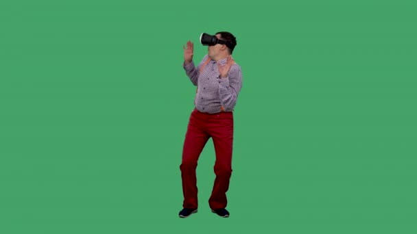 Porträtt av en man med en virtuell verklighet headset eller 3D-glasögon på huvudet. En man med glasögon, en skjorta med orange hängslen i studion på en grön skärm. Långsamma rörelser. — Stockvideo