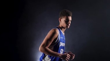 Stüdyoda siyah bir arka planda sahne ışığı altında poz veren basketbol oyuncusu üniformalı genç bir adam. Adam kendine güvenen bir basket topu yakalıyor, top sürüyor. Ağır çekim. Kapat..