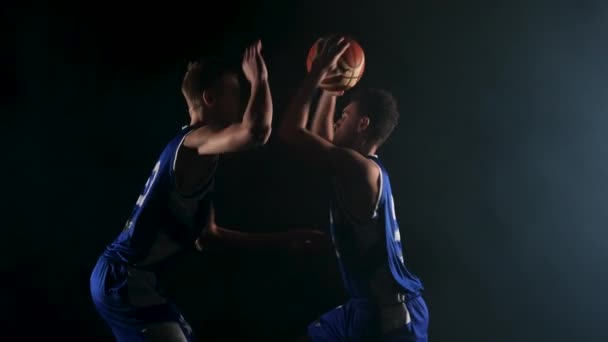 Due ragazzi stanno lottando per una palla da basket. Giovani giocatori di basket si allenano in uno studio buio fumoso su uno sfondo nero. Al rallentatore. Da vicino.. — Video Stock