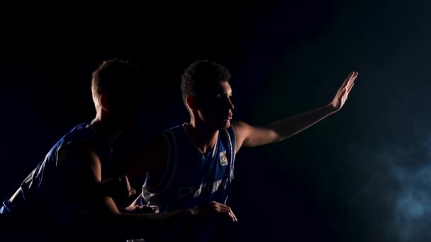 Молодые спортсмены-баскетболисты тренируются в темно-дымчатой студии на черном фоне. Афроамериканец ловит мяч и обходит противника. Медленное движение. Закрыть. — стоковое видео