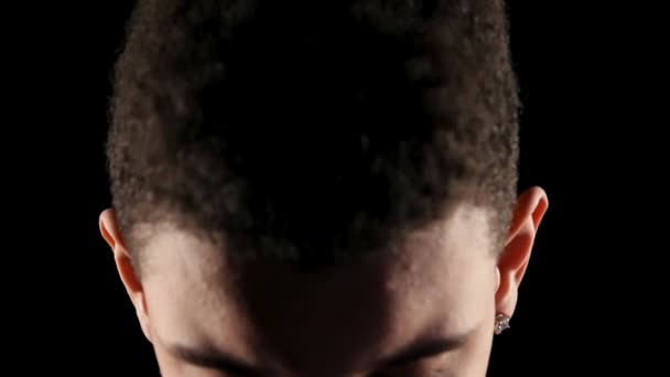 Retrato de un joven jugador de baloncesto afroamericano en un estudio oscuro sobre un fondo negro. El tipo levanta la cabeza inclinada y mira hacia adelante en cámara lenta. Rostro cercano de un joven. — Vídeos de Stock