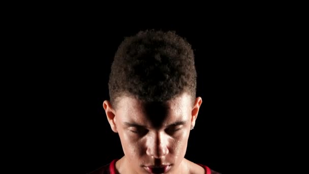 Portrait of a young African American basketball player in a dark studio on a black background. The guy raises his bowed head and stares straight ahead in slow motion. Close up. — Stock Video