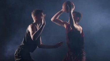 İki genç sporcu, spot ışıkları altında karanlık dumanlı bir stüdyoda basketbol antrenmanı yapıyor. Afrikalı Amerikalı topu yakalıyor ve rakibini geçmeye çalışıyor. Ağır çekim. Kapat..