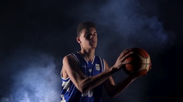 A guy catches the ball, dribbles it behind his back, and throws it. Portrait of a basketball player in a smoky studio on a black background in the light of a searchlight. Slow motion. Close up. — Stock Video