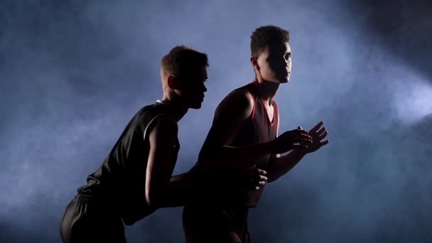 Dois jovens atletas praticam basquete em um estúdio fumegante escuro sob os holofotes. O afro-americano apanha a bola e dá a volta ao adversário. Movimento lento. Fechar. — Vídeo de Stock