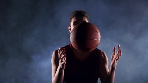 Porträtt av en ung afroamerikansk basketspelare i en mörk rökig studio i rampljuset. Killen kastar en basketboll, snurrar den. Silhuett. Sakta i backarna. Närbild. — Stockvideo